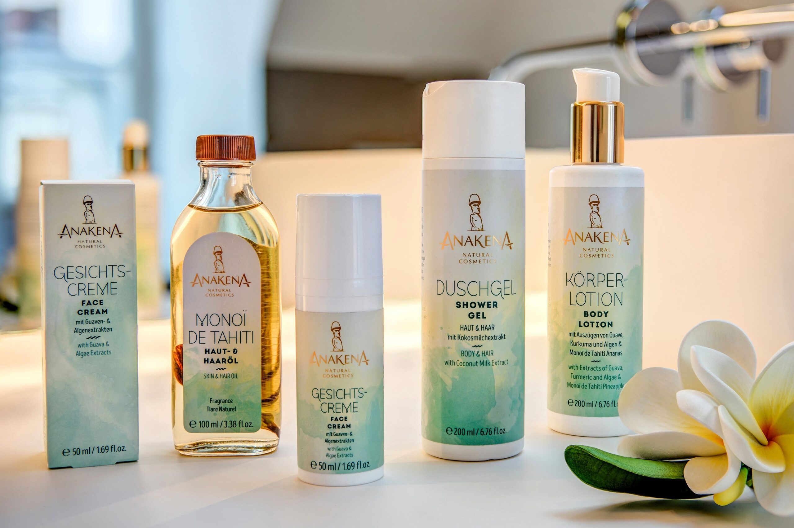 Produkte von Anakena - Natural Cosmetics mit Gesichtscreme, Monoï de Tahiti, Duschgel Haut & Haar und Körperlotion, arrangiert mit Tiaré-Blüten.
